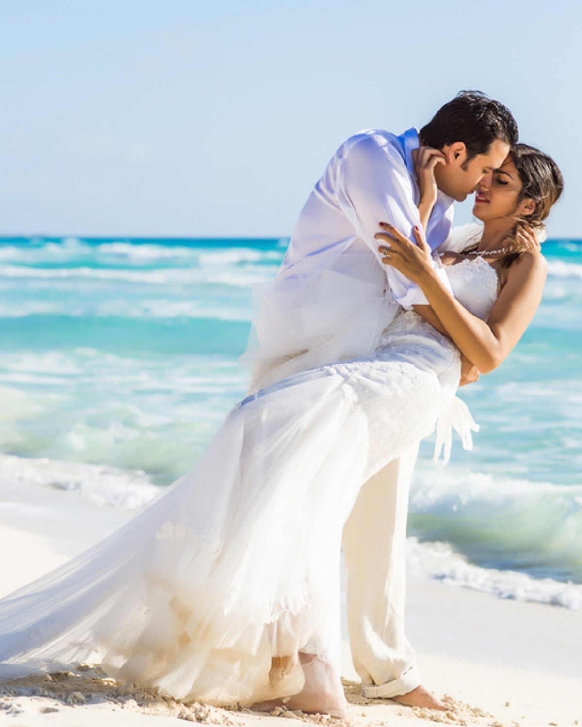 A newlywed couple kissing on the beach, the bride in her wedding dress with a flowing train