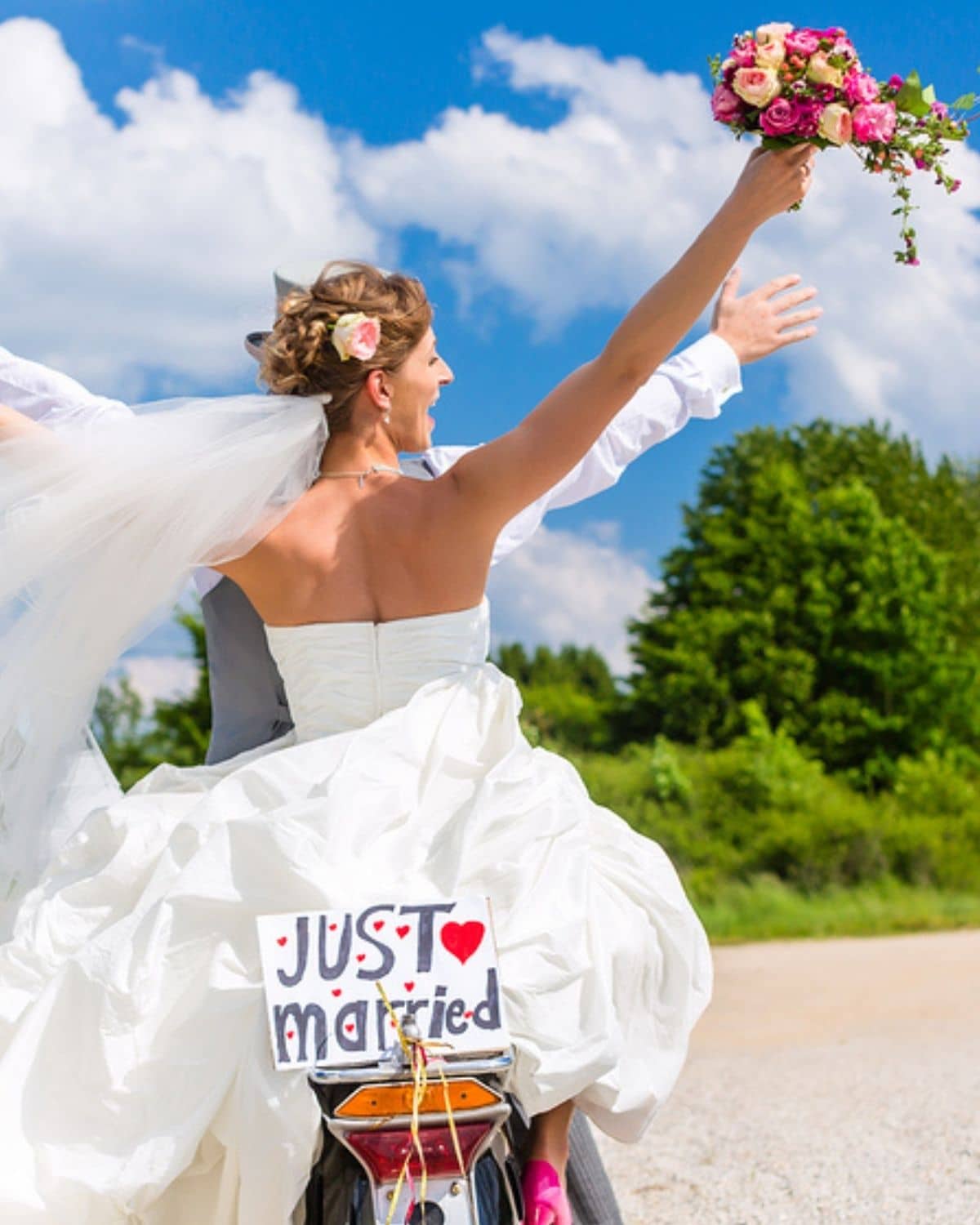 A just-married bride in her wedding dress waves her bouquet from the back of a 'Just Married' scooter, veil flowing in the sun
